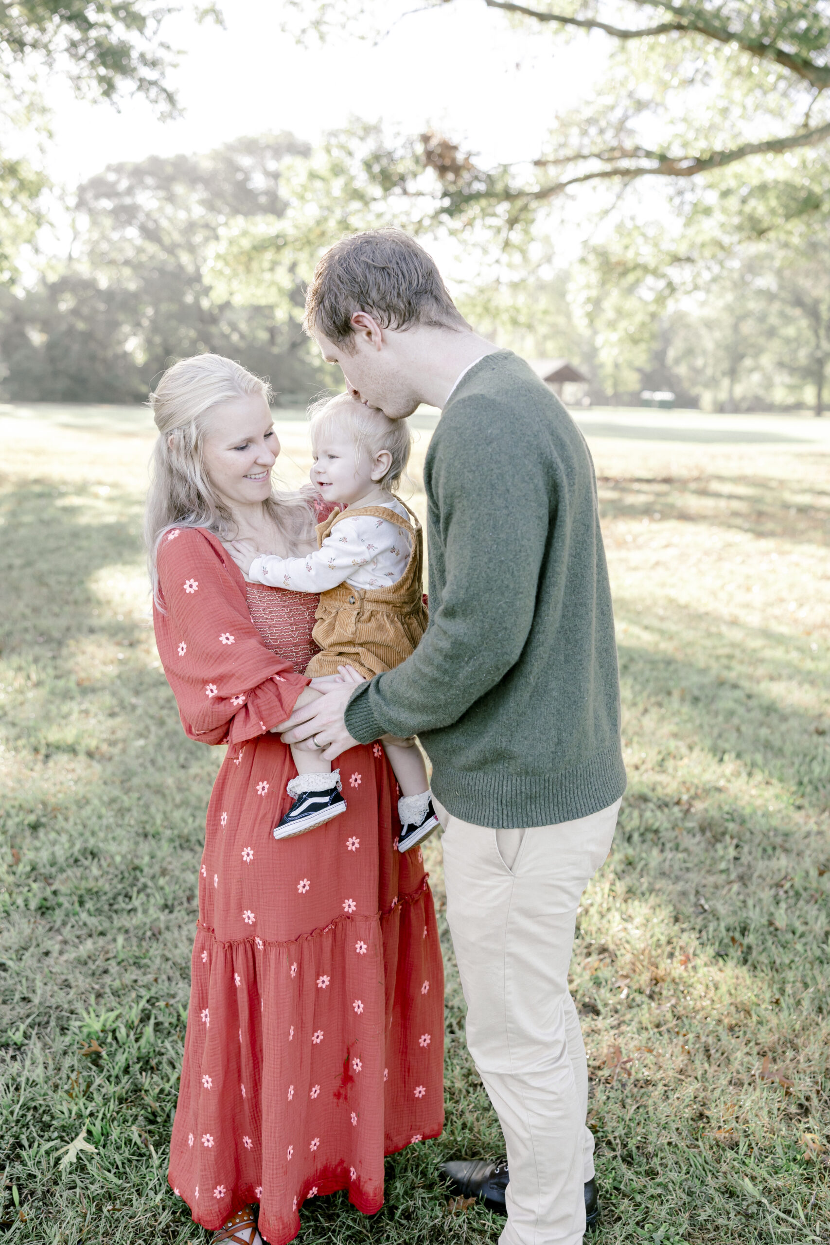 Mom and dad holding toddler in Fort Hunt Park. Dad is kissing the toddler's head.