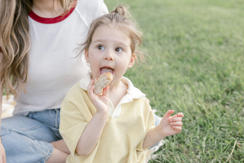 A toddler girl sitting on a picnic blanket eating a piece of bread during a relaxed park family photo session in Northern Virginia.