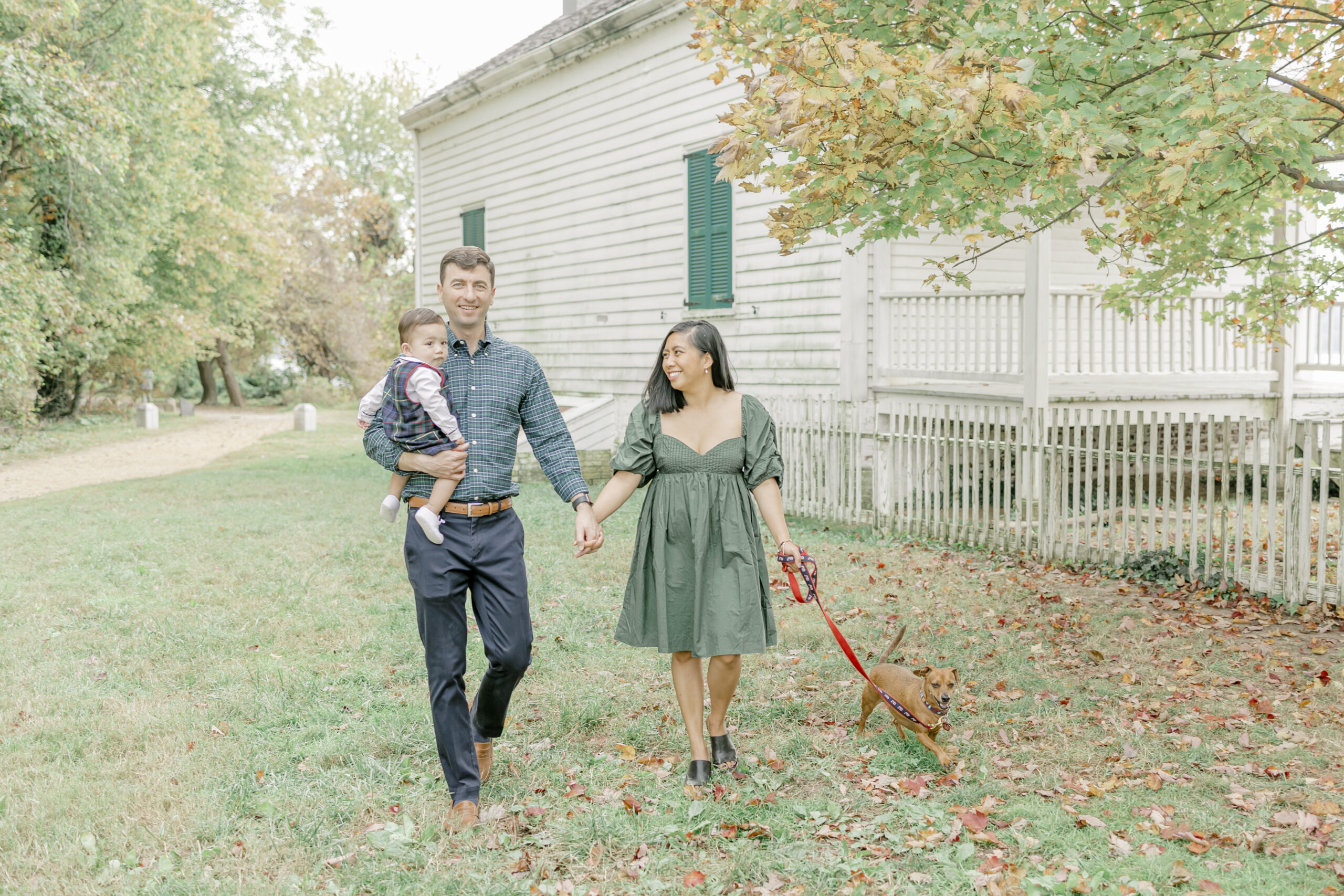 A man and woman walk hand-in-hand, with the father holding their toddler and the mother walking their dog. Light autumn foliage and a white building provide a serene backdrop for fall family photos