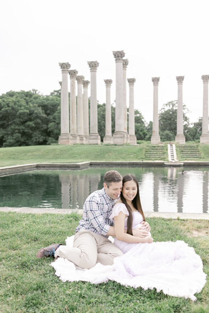 Man and woman sitting on a blanket outdoors, smiling and looking down at the woman’s pregnant belly.