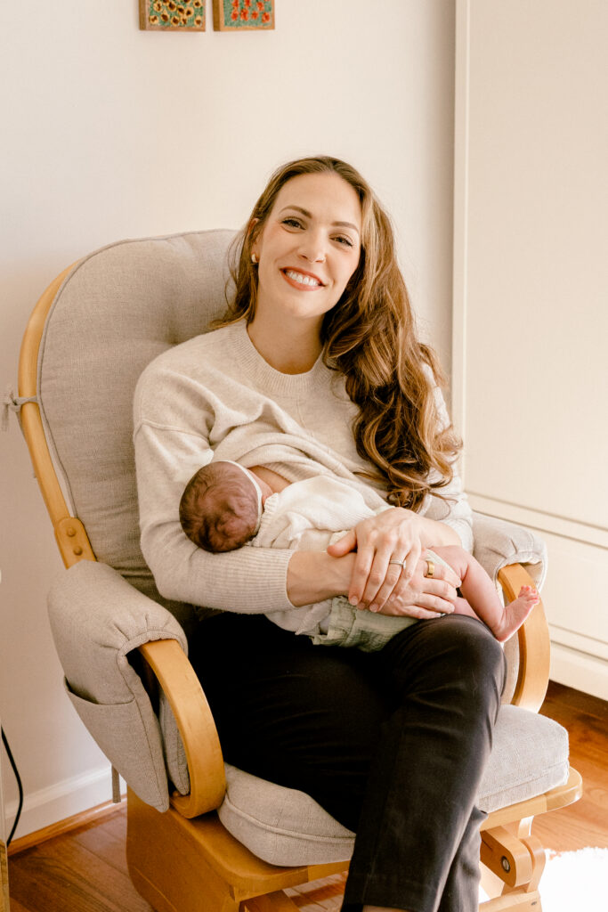 Mother smiling while breastfeeding her newborn baby in a softly lit in-home postpartum setting.